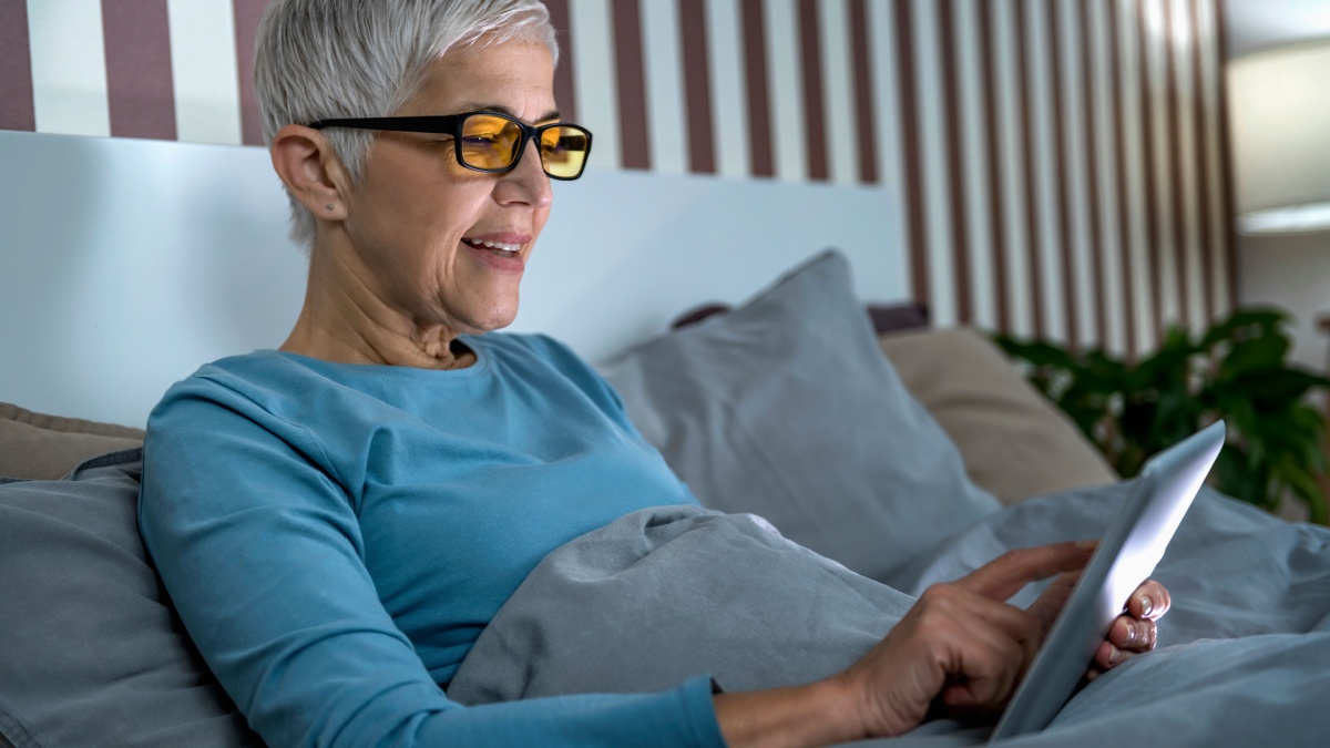 Mature woman wearing blue light blocking glasses with amber lenses, lying in bed before sleep, looking at tablet screen.