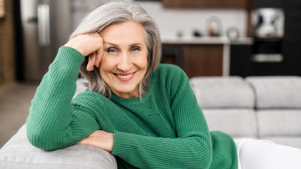 Portrait of charming senior woman with grey hair, smiling mature female sits on the couch at ease and looks at the camera.