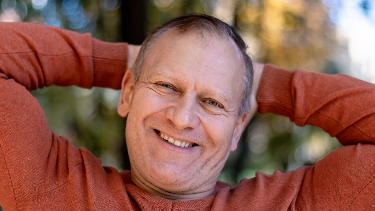 portrait of a man, handsome smiling, charming middle age, 55-60 years old, with gray hair, with wrinkles, in a red sweater, holding his hands behind his head, against the background of nature.