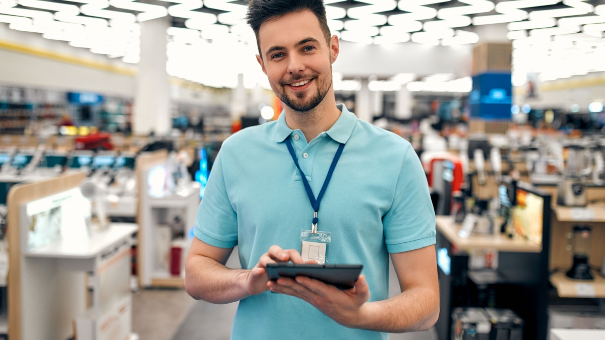 A young successful sales assistant in uniform with a tablet in his hands against the background of the interior of the hall in a home appliances and electronics store.