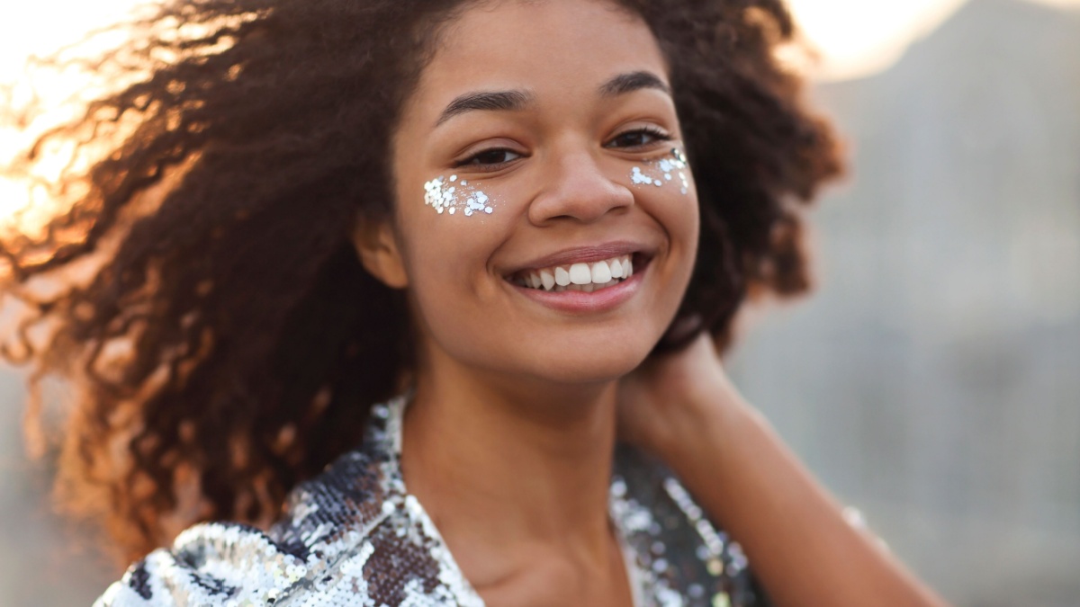 Close up portrait of overjoyed charming african american woman with curly hair and glitter on cheekbones smiling happily at camera while moving dancing outdoors, festive atmosphere in the air.