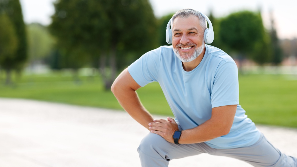 Happy positive mature sportsman during outdoor workout, senior man wearing headphones and sports outfit warming up muscles, doing side squat on one leg, enjoying active lifestyle outside in park.