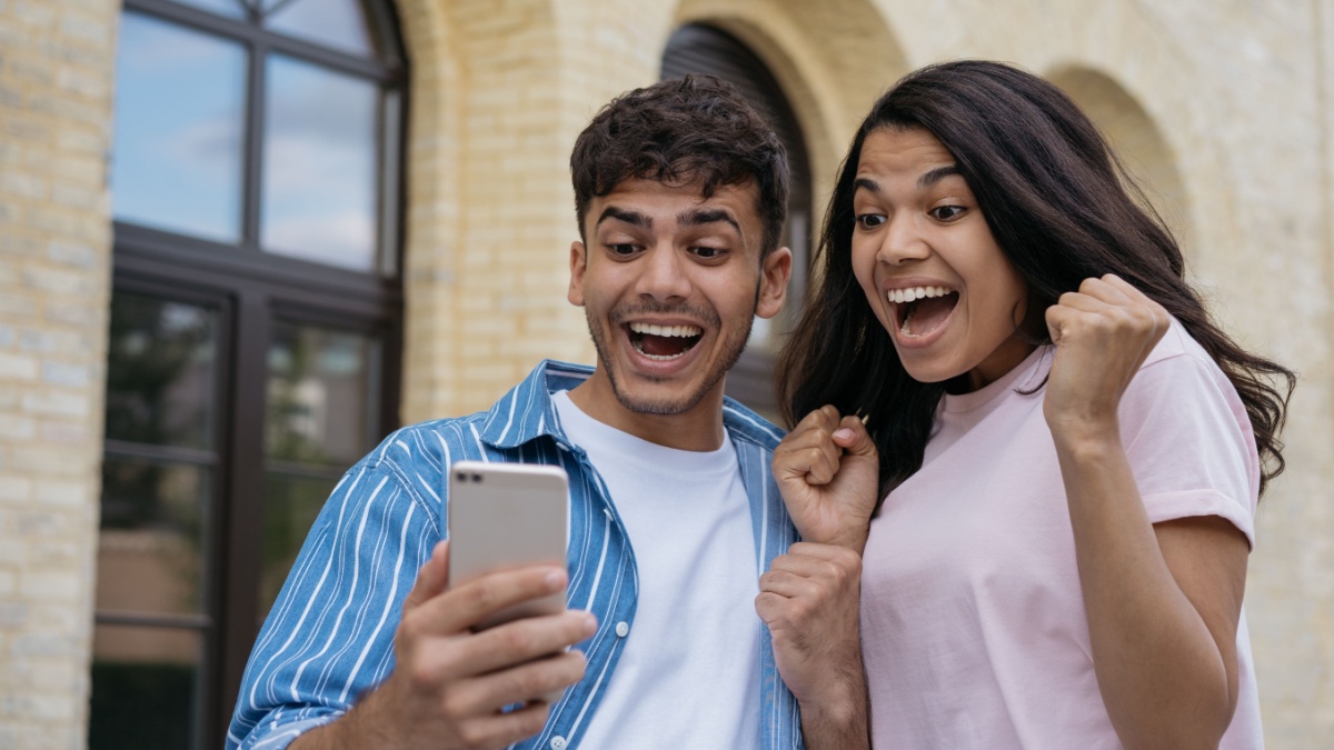 Overjoyed couple of friends using smartphone playing mobile game, celebration success. Emotional Indian man and African American woman shopping online with sales. Excited freelancers receive payment.