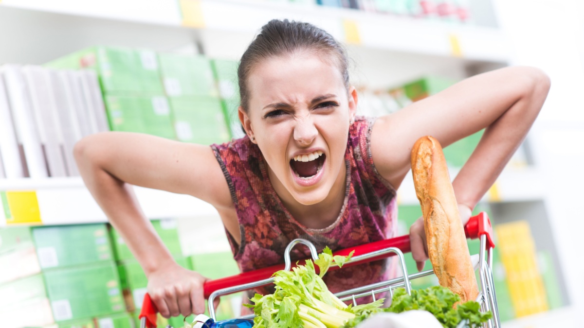 Angry woman pushing a full shopping cart at store with shelves on background.