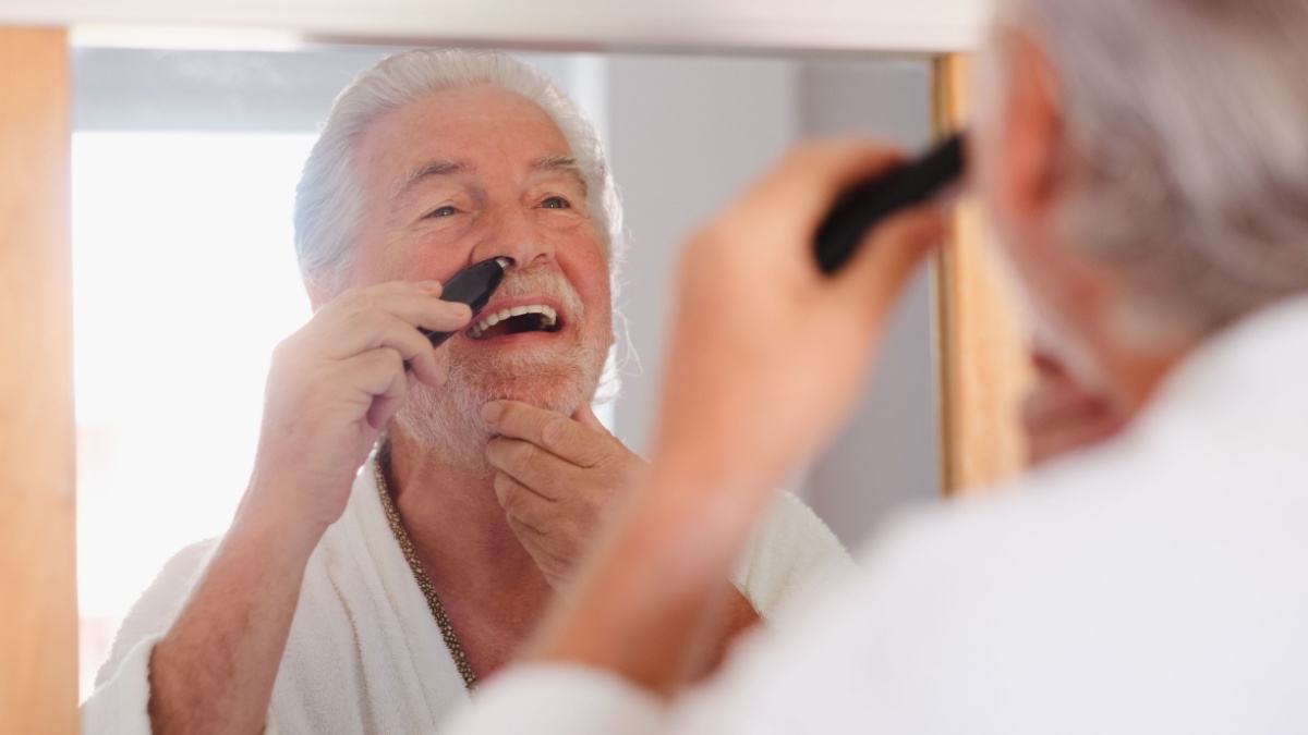 Portrait of senior bearded man in bathrobe while remove nose ear hair with help of little comfy trimmer looking at himself in the mirror