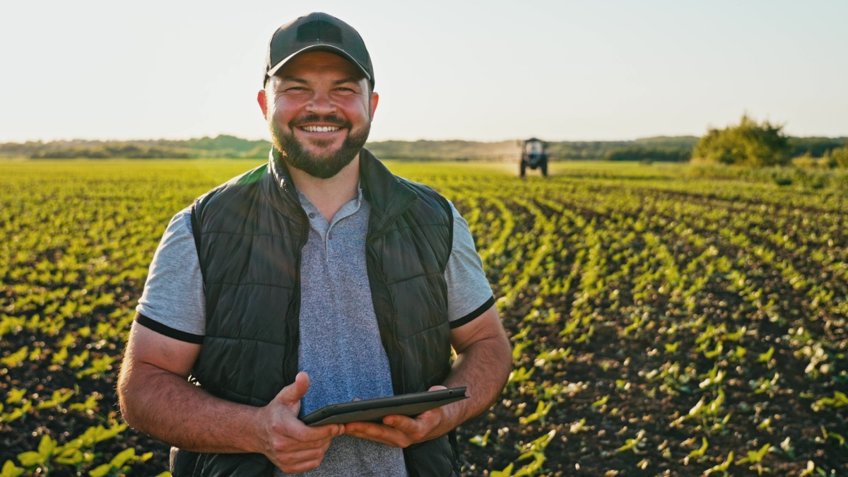 Happy farmer stands and smile holds tablet in his hands against background of working tractor in field.