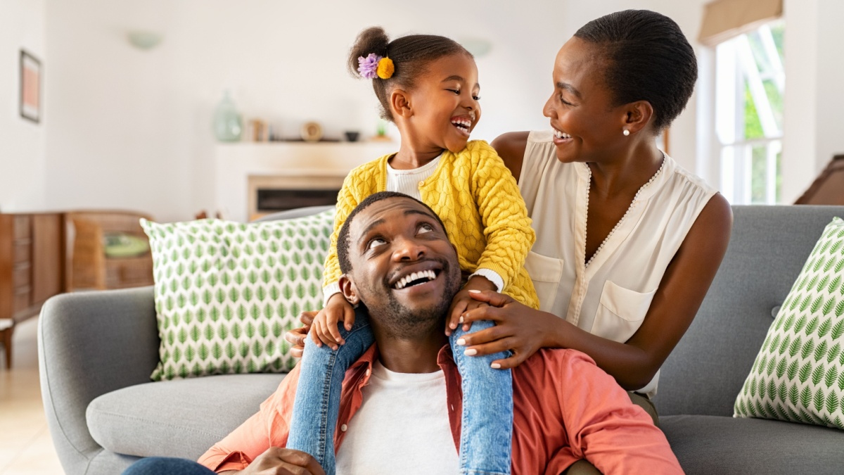 Cheerful little girl sitting on father shoulder while playing with mother at home. Happy black family enjoying weekend at home. Cute little daughter sitting on fathers shoulder and play with her mom.
