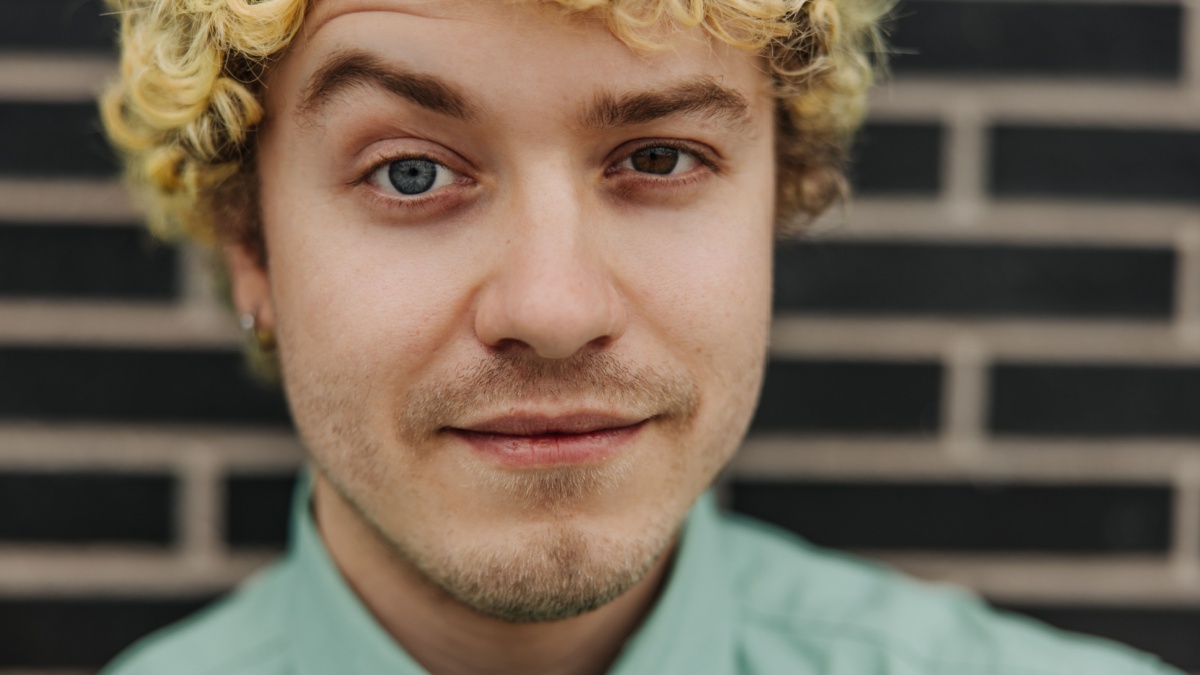Close-up of young caucasian guy casually smiling at camera with raised eyebrow. Blond-haired man with black wall in background.
