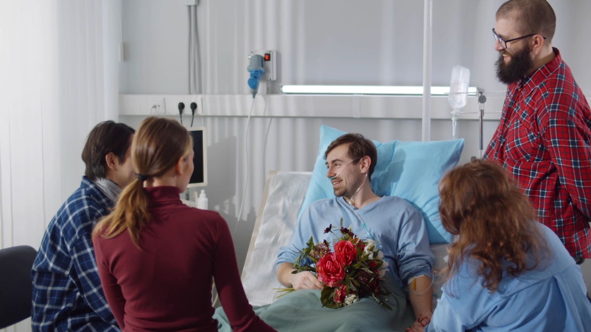 Young people visiting friend in hospital and bringing flowers. Smiling male patient lying in hospital bed and talking to friends during visit in clinic room
