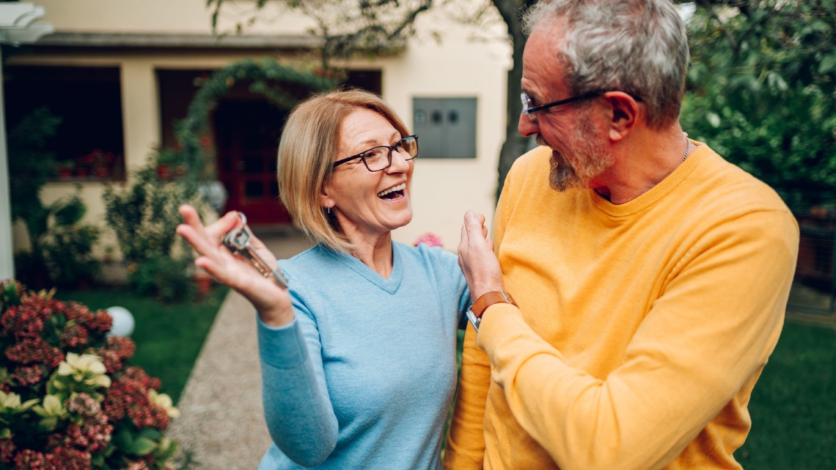 Portrait of a happy mature couple bought their new home and standing in front of it and showing keys. Real estate owners. Senior married couple posing in front of their new family house.