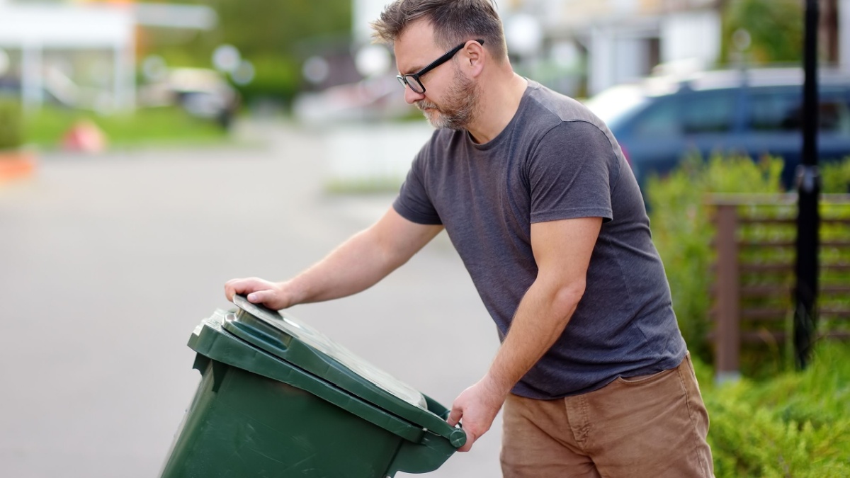 Mature man pulling out a large green plastic garbage container in front of the townhouse to the roadway of the street.