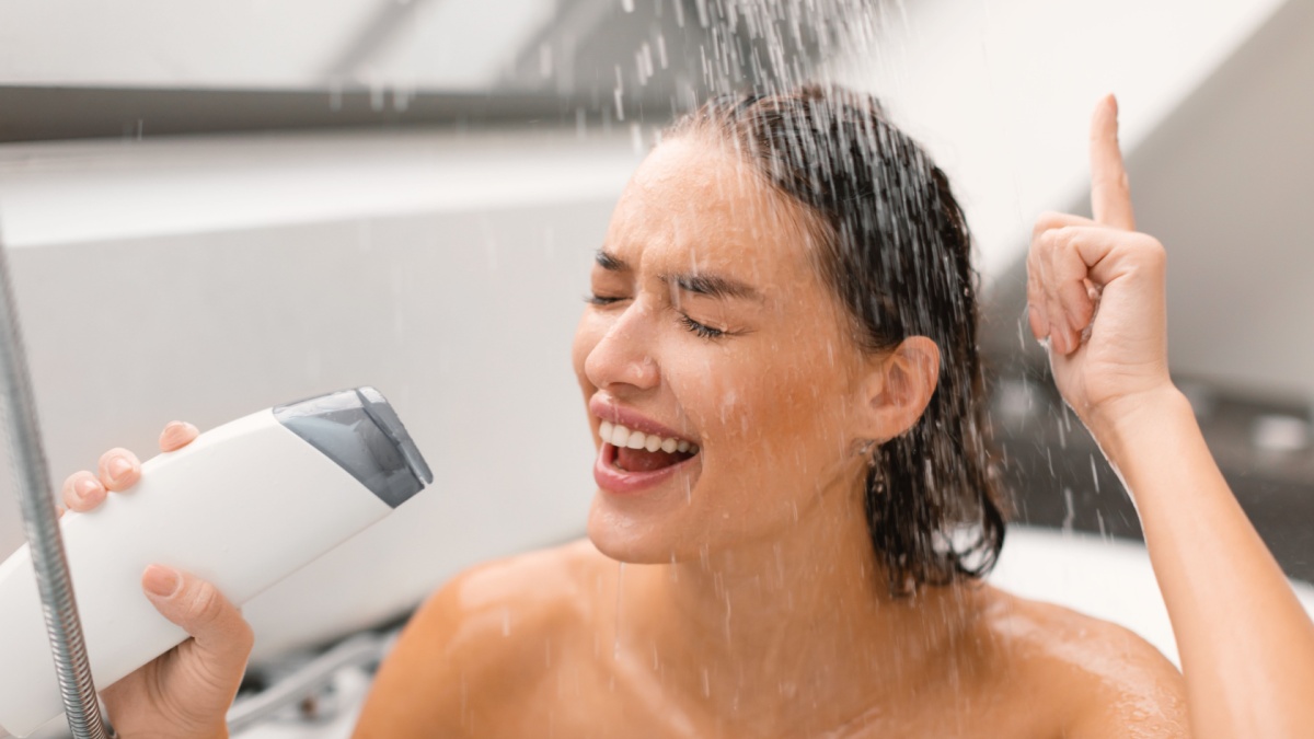 Cheerful Female Taking Shower Singing Holding Shampoo Bottle Like Microphone, Enjoying Body And Hair Care Routine Sitting With Eyes Closed In Modern Bathroom Indoors.