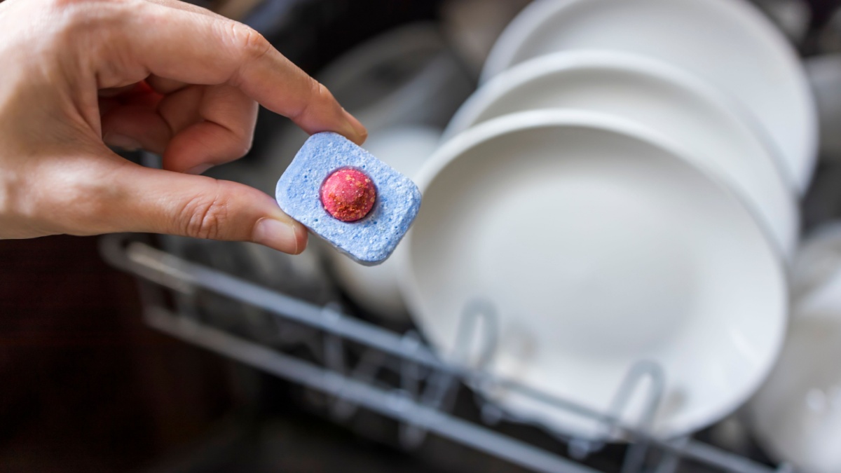 Close up of woman hand filling dishwasher tablet into open automatic stainless built-in dishwasher machine with dirty white dish inside in modern home kitchen.Household, housekeeping domestic life.