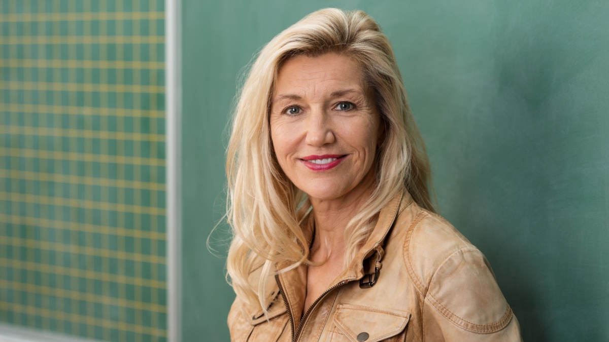 Attractive stylish middle-aged woman teacher standing in front of the class blackboard looking thoughtfully at the camera with a smile.