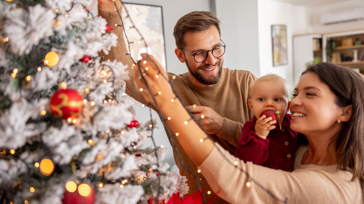 Young parents having fun decorating Christmas tree with their cute little baby girl, placing Christmas lights on it while decorating home for winter holiday season
