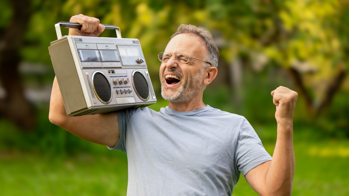 Happy excited middle aged man listening to retro tape recorder and dancing and joyfully gestured to his favorite disco song outdoors in park in summer sunny day. He remembers his youth.