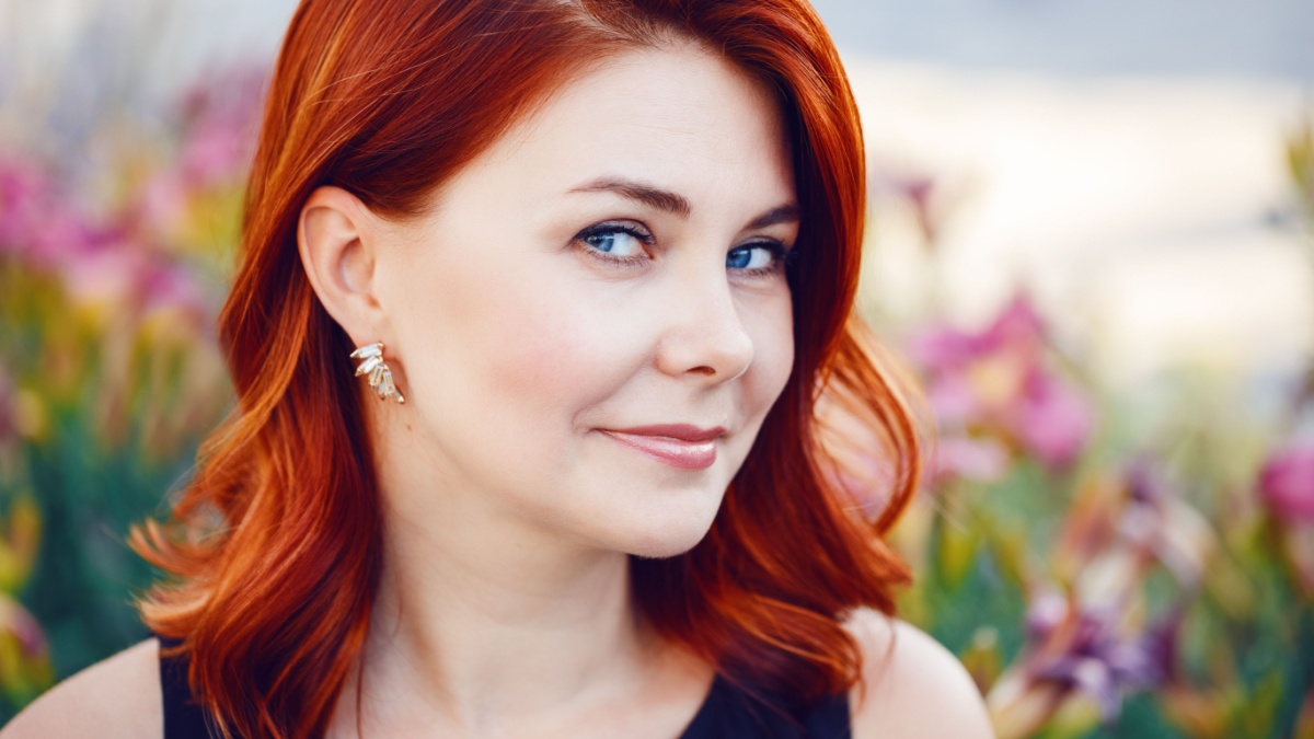 Closeup portrait of smiling middle aged white caucasian woman with waved curly red hair in black dress looking in camera outside in park among flowers, beauty fashion lifestyle concept.