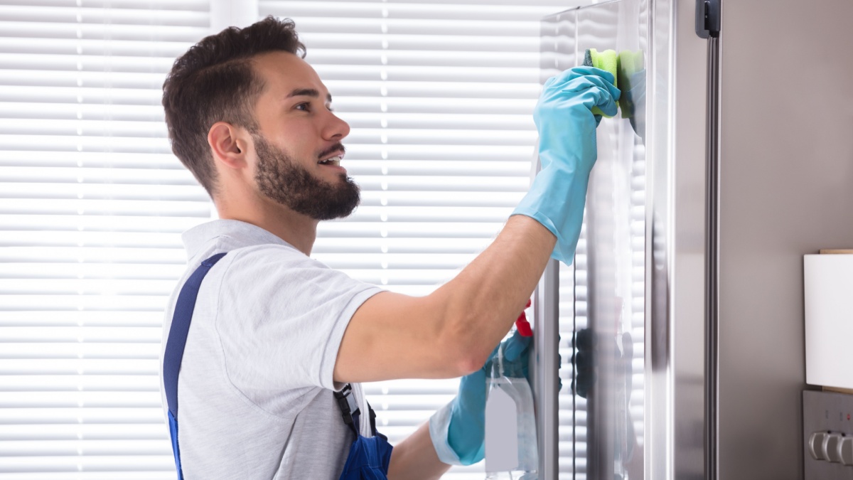 Side View Of A Young Male Janitor Cleaning Refrigerator In Kitchen