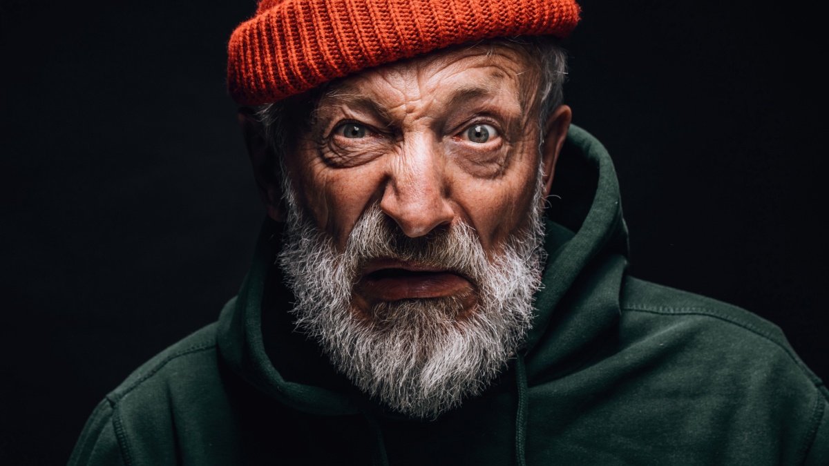 Portrait of old weird bum in orange hat with crazy eyes and puzzled confused expression on black studio background.