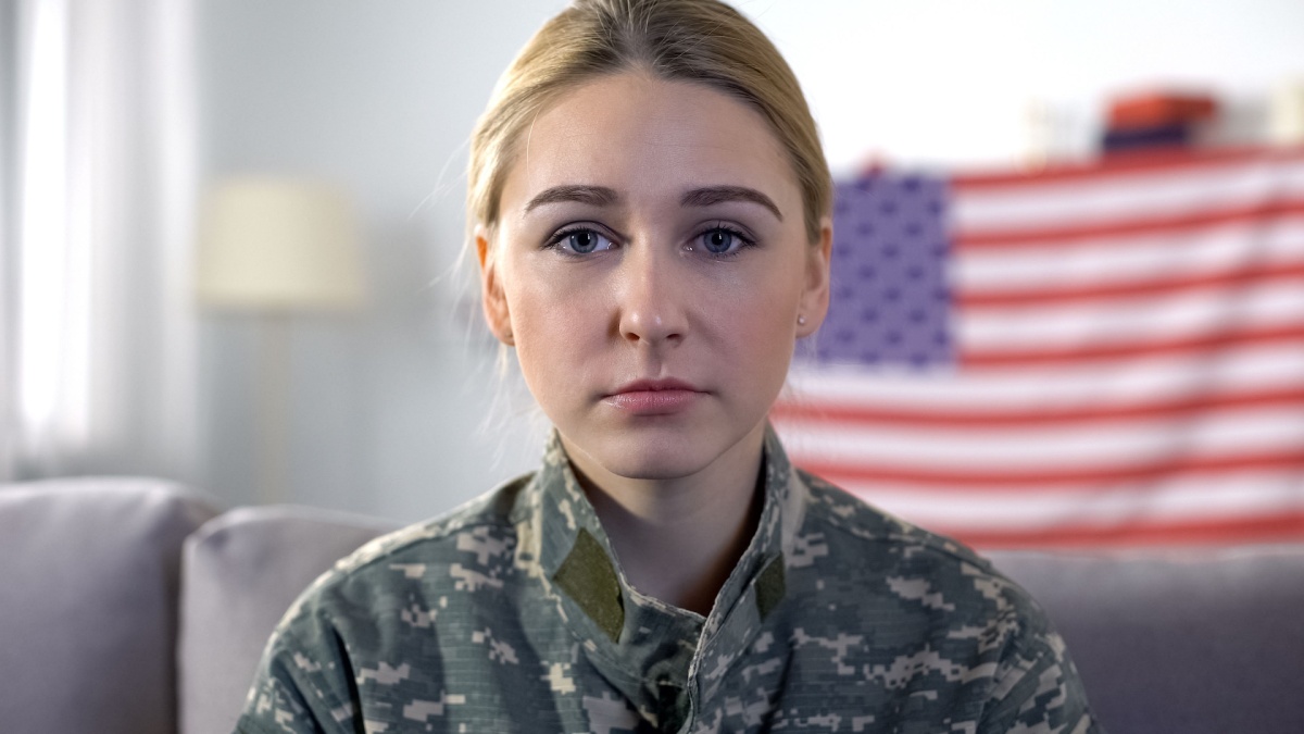 Sad female soldier in camouflage uniform looking at camera, USA flag background.