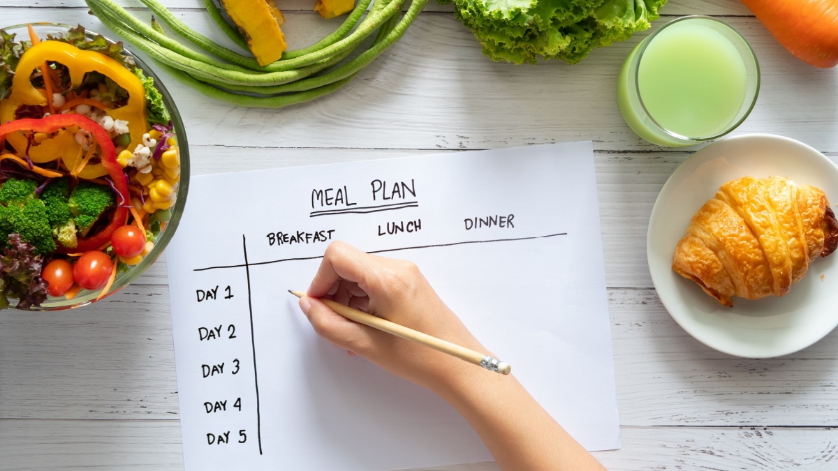 Calories control, meal plan, food diet and weight loss concept. top view of hand filling meal plan on weekly table with salad and fresh vegetable on dining table.