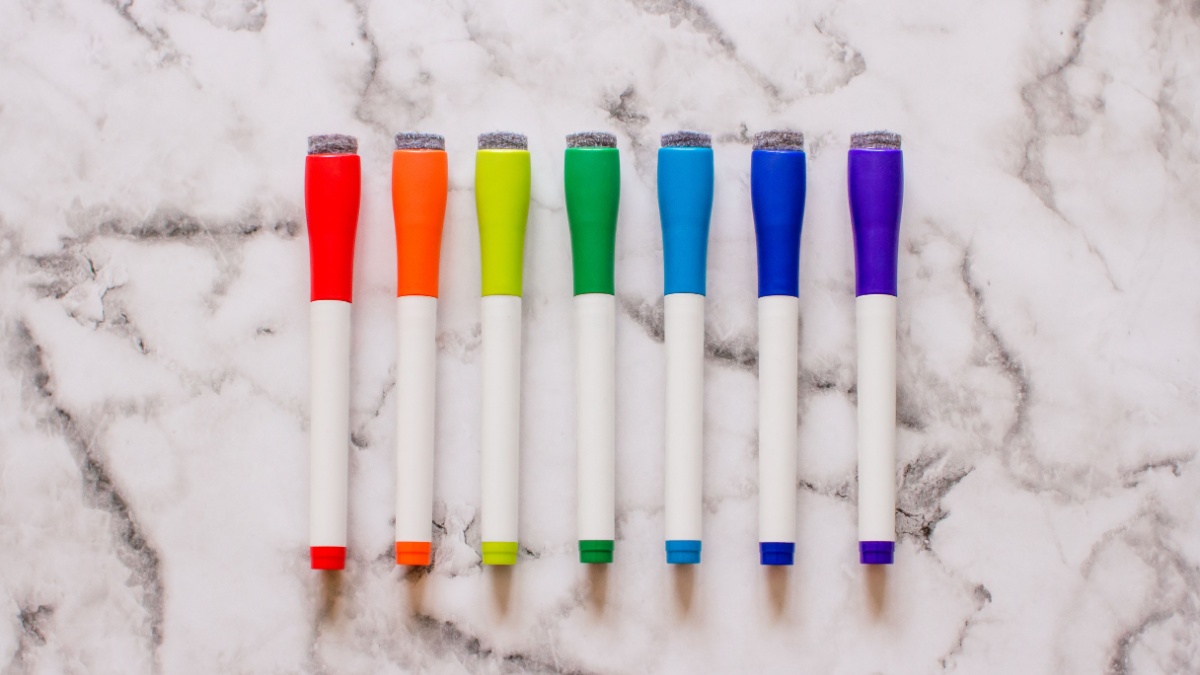 Top down view of dry erase markers lined up in a row and organized in rainbow order on top of a marble surface