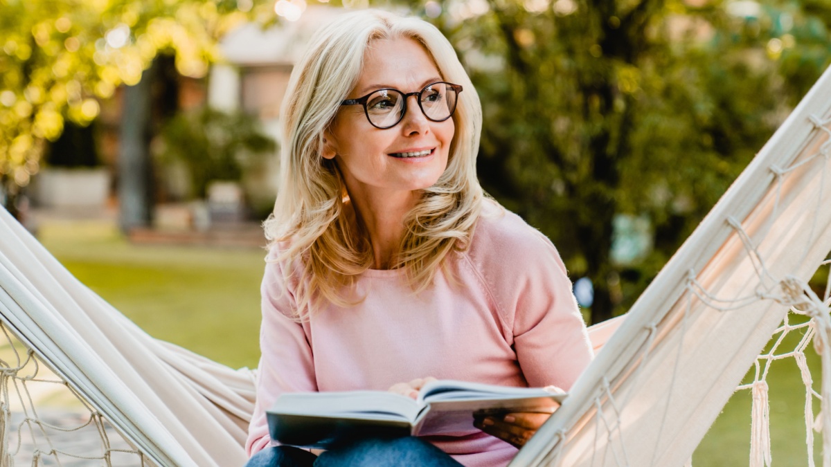 Smiling senior good-looking blond woman wearing glasses while reading in hammock in the summer garden.