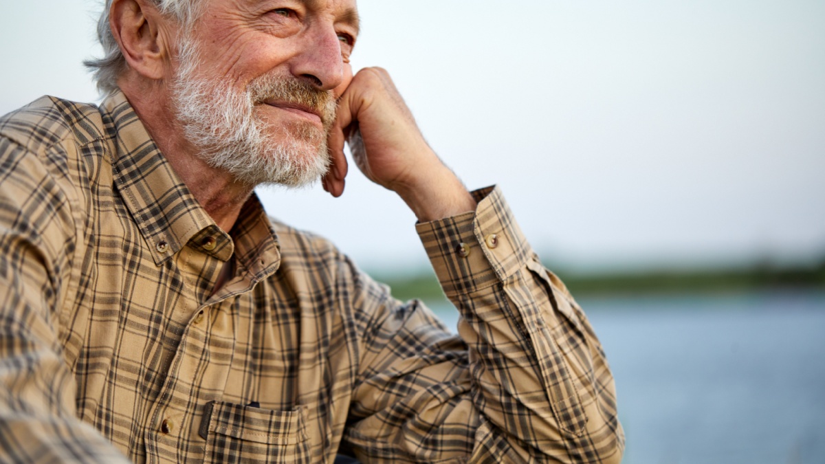 Pleasant Senior Man In Checkered Shirt Sit On Chair In Nature, Having Rest Alone, Thinking, Smiling, In Contemplation Of Countryside Landscapes, Portrait Of Gray Bearded Male Relaxed.