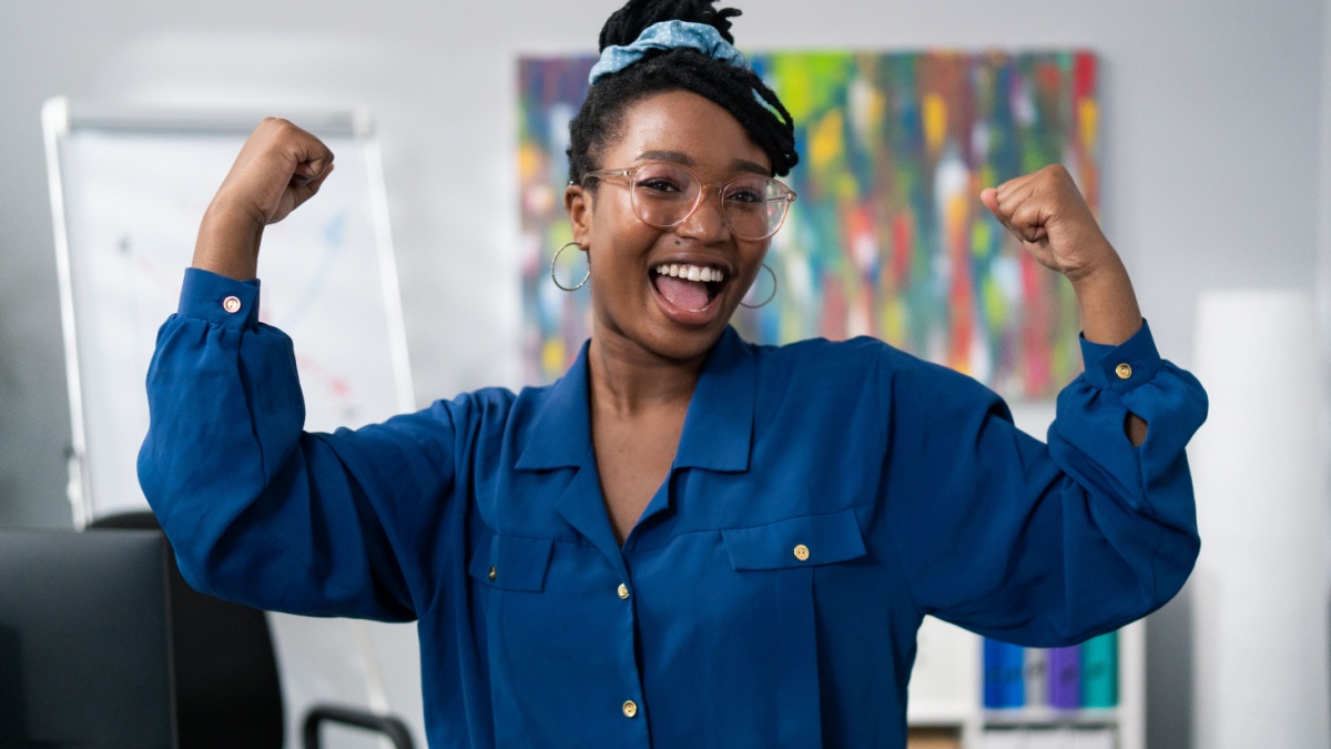 Dark-skinned woman wearing blue clothes glasses stands against office backdrop smiles happy, victory, winning, raises hands up tenses hand muscles.