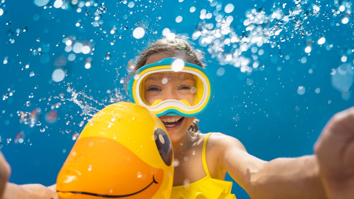 Happy child having fun on summer vacation. Funny kid jumping in swimming pool. Low angle view portrait of girl against water splash. Spring break!