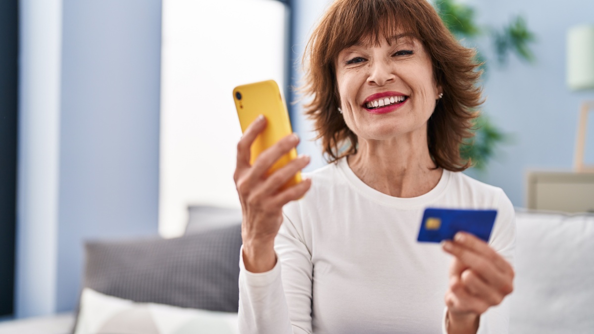 Middle age woman using smartphone and credit card sitting on sofa at home.