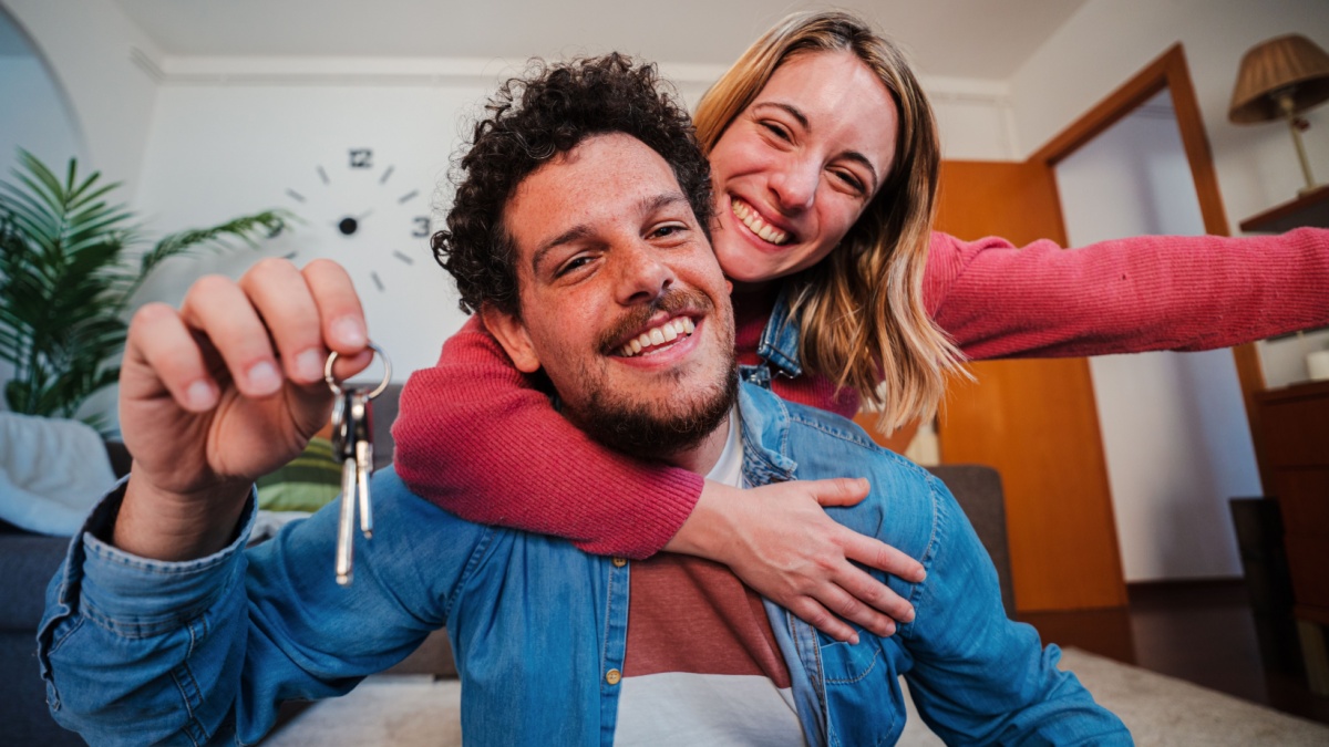 Young caucasian couple showing keys of their first house after purchase and moving to new home together. happy husband and wife hugging in their apartment excited to be owners of a apartment.