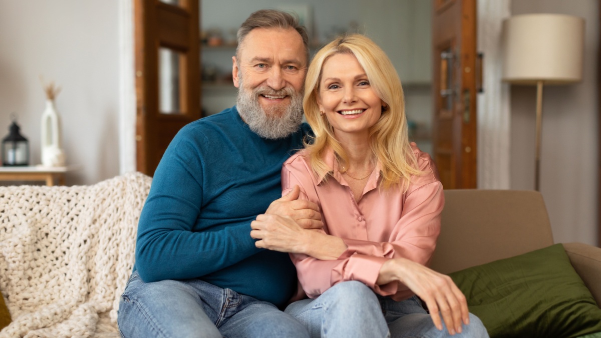 Love In Mature Age. Happy European Couple Embracing Sitting On Couch Smiling To Camera At Home.