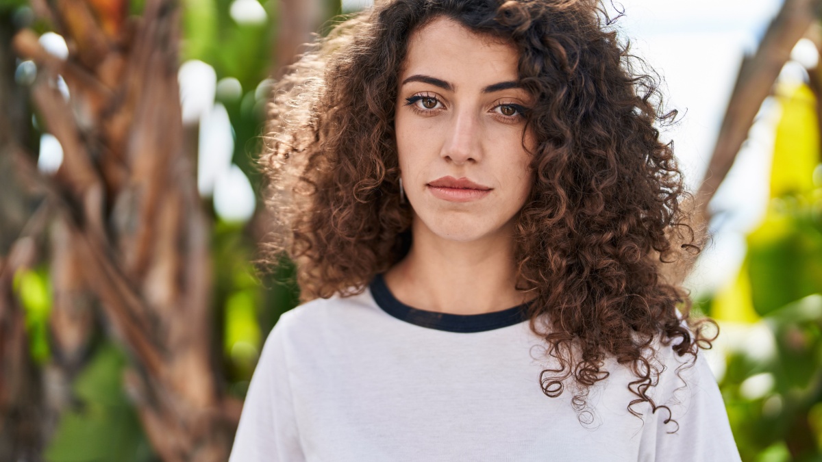 Hispanic woman with curly hair standing outdoors thinking attitude and sober expression looking self confident.