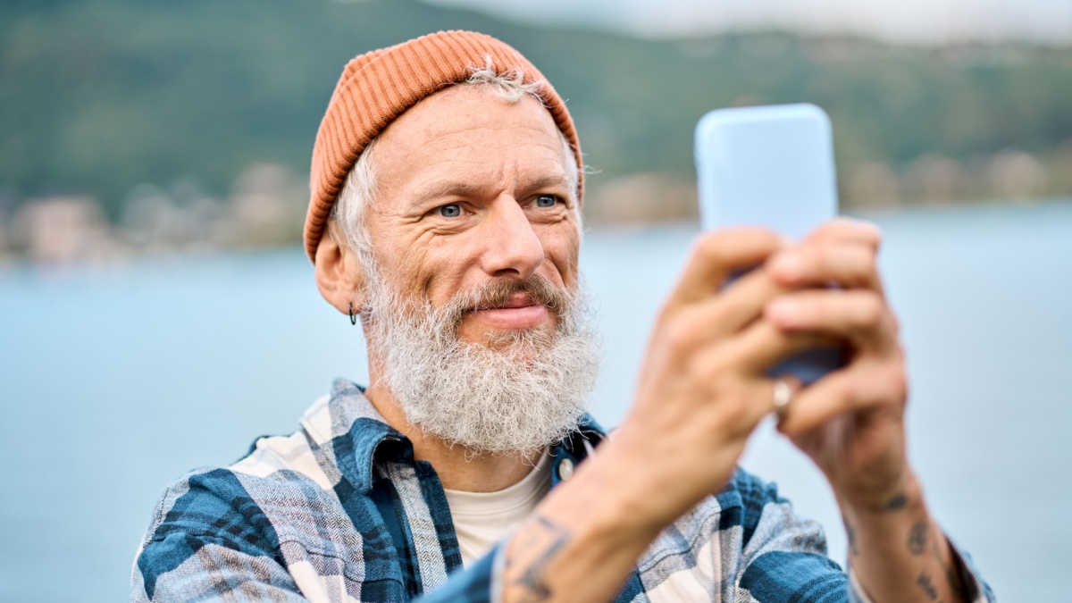 Cool bearded happy older elder hipster man standing in nature park taking photos on smartphone using mobile phone. Mature boomer traveler enjoying.