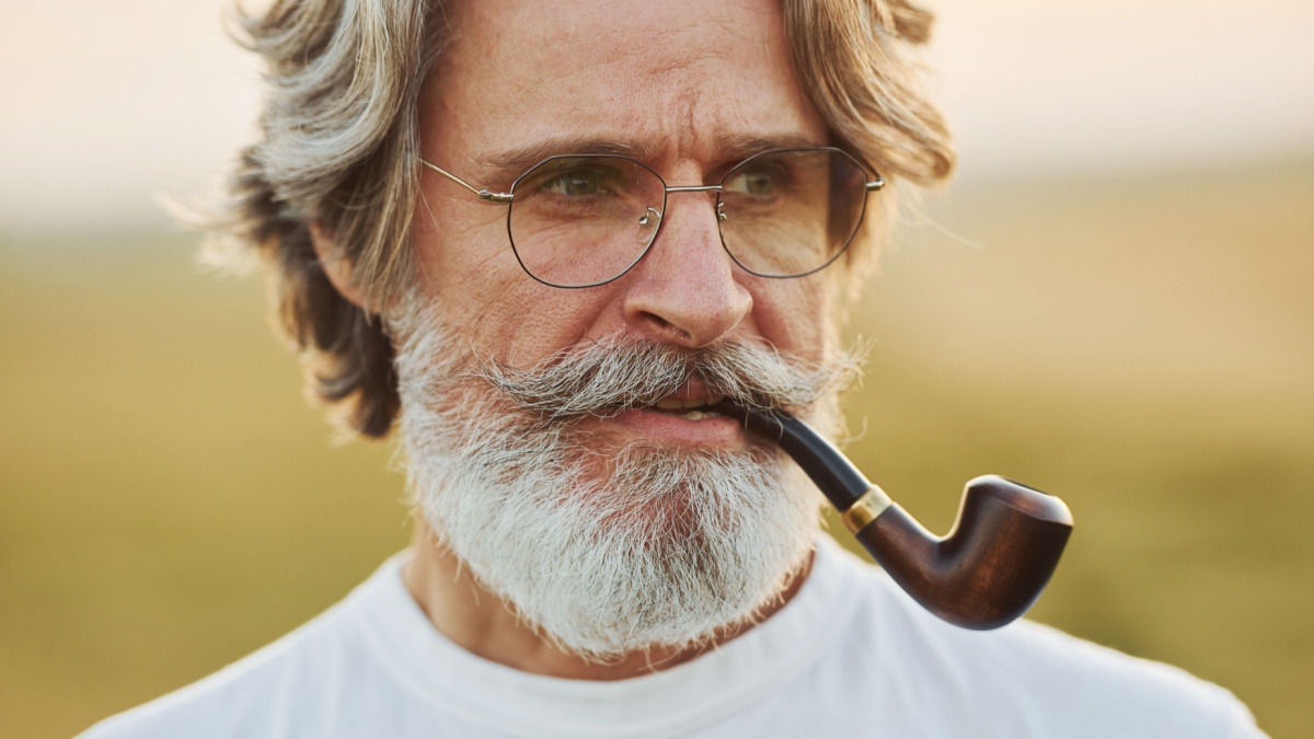 Portrait of senior stylish man with grey hair and beard that standing outdoors on field at sunny day and smoking.