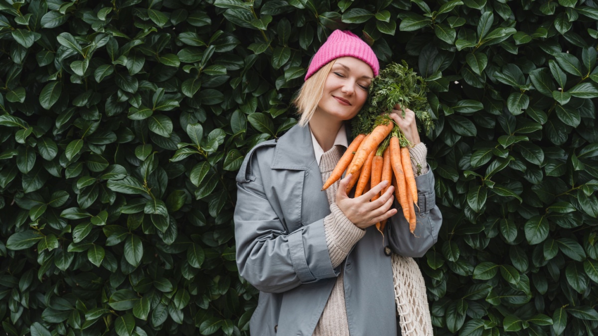Smiling woman with fresh organic carrots and mesh bag against green leaf wall. Concept of people doing sustainable shopping of eco local products without plastic packaging. Minimalist lifestyle