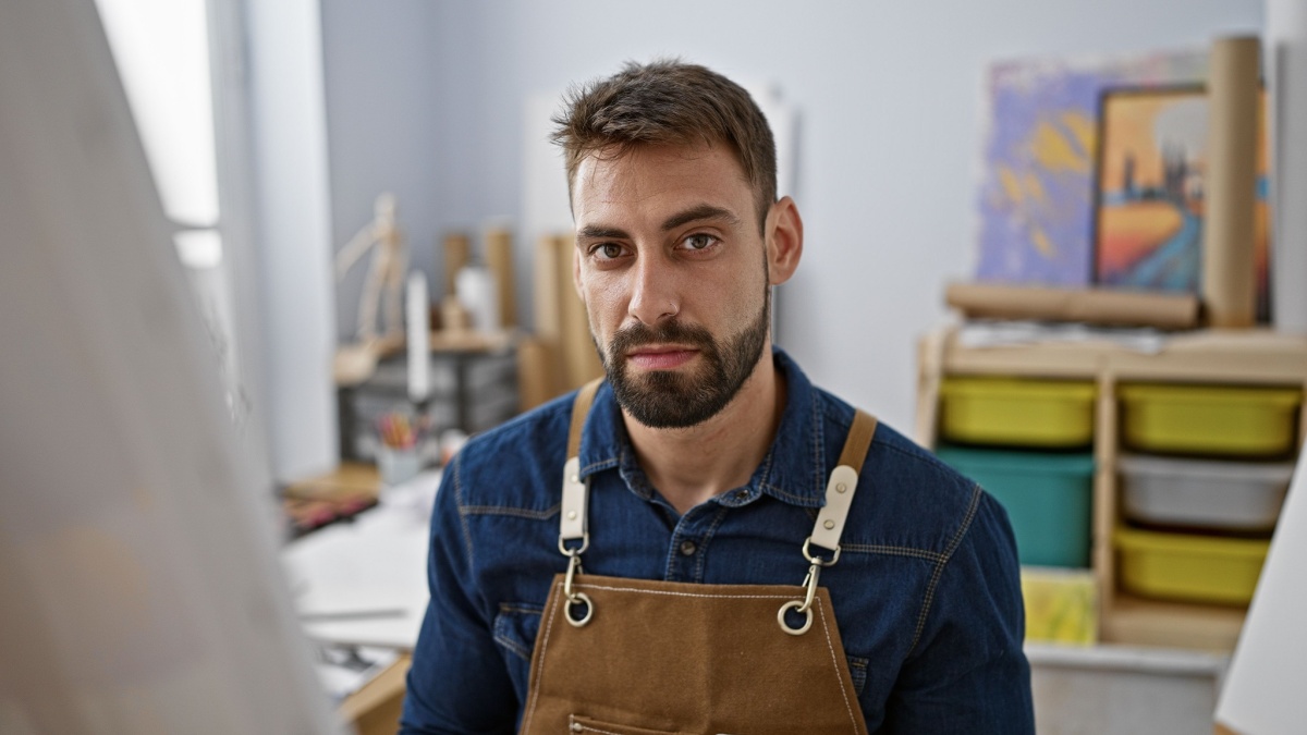 Focused young hispanic male artist sitting inside art studio, handsome bearded man engaged in serious brush-to-canvas painting.