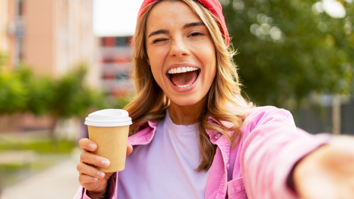 Beautiful smiling woman wearing red stylish hat holding cup of coffee taking selfie on the street.