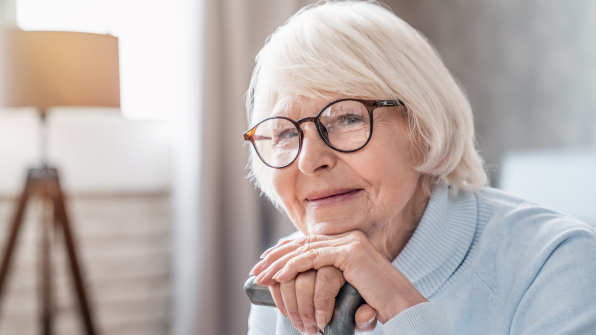 Close up of mature woman in glasses holding cane while sitting on sofa at home. Loneliness and calmness, nostalgy concept. Hospice nursing house.