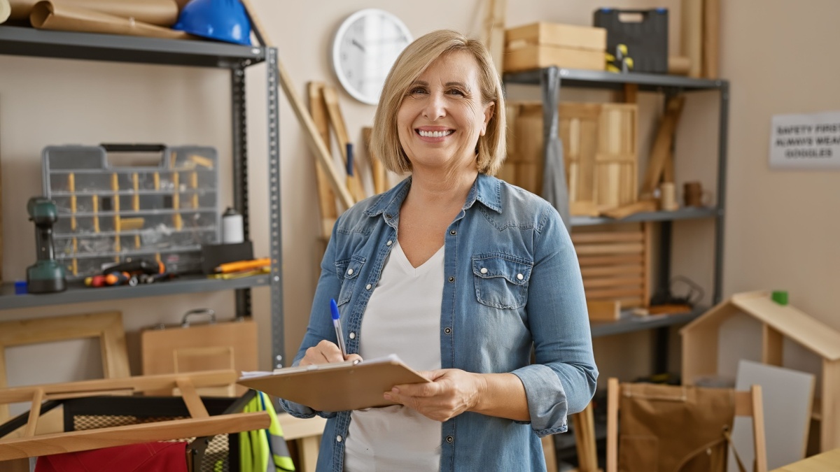 Smiling mature woman with clipboard standing in a well-organized carpentry workshop.