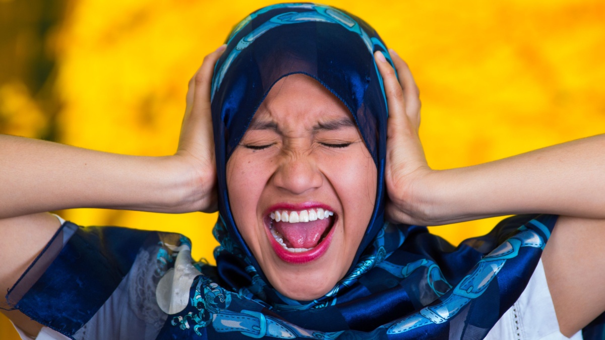 Beautiful young muslim woman wearing blue colored hijab, facing camera covering ears with hands screaming out, autumn forest background.