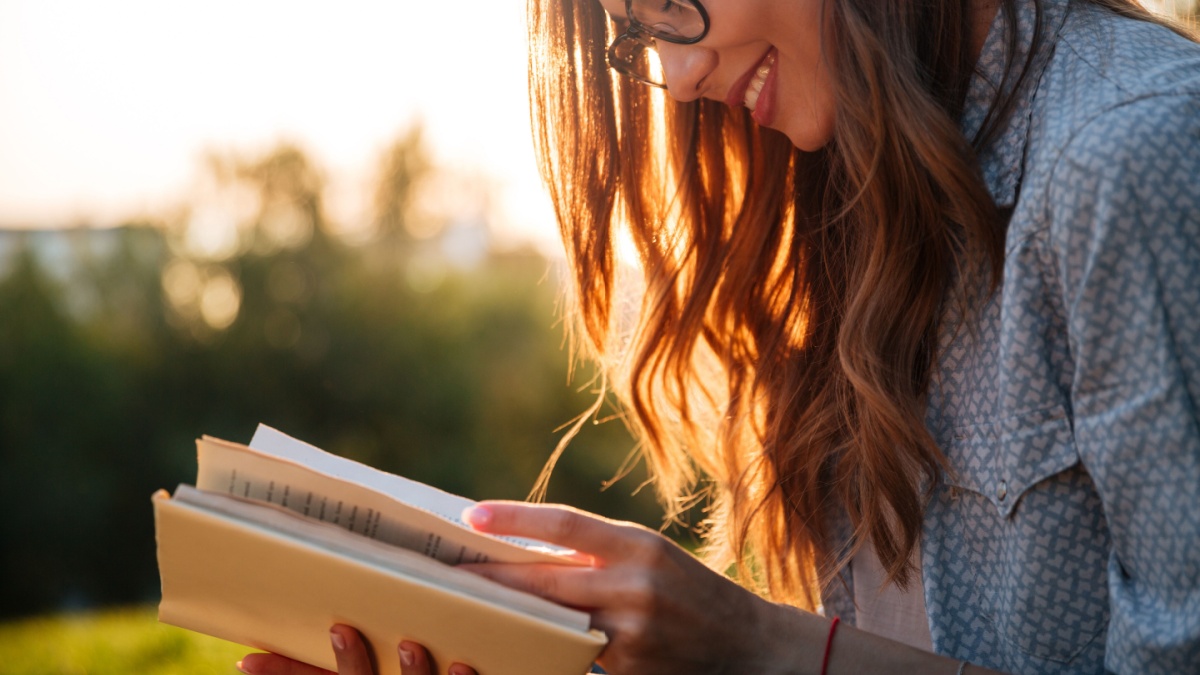 Cropped image of smiling brunette woman in eyeglasses reading book in park.