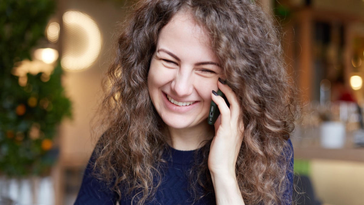Cheerful excited young European female with curly gorgeous hair wearing blue sweater laughing with tender as she receives good positive news.