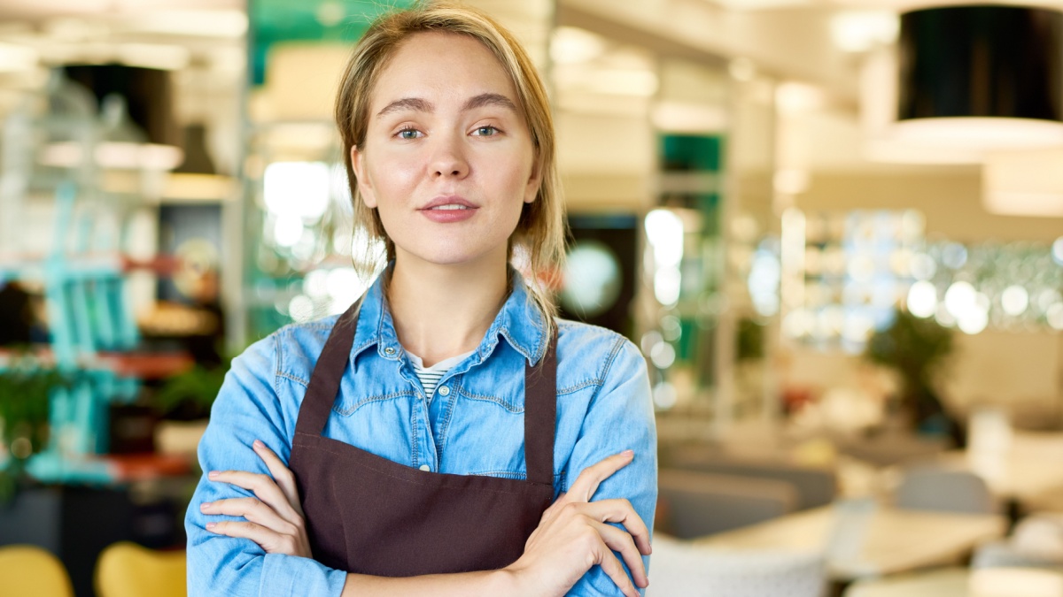 Waist up portrait of young waitress wearing apron posing confidently with arms crossed and looking at camera in cafe interior.