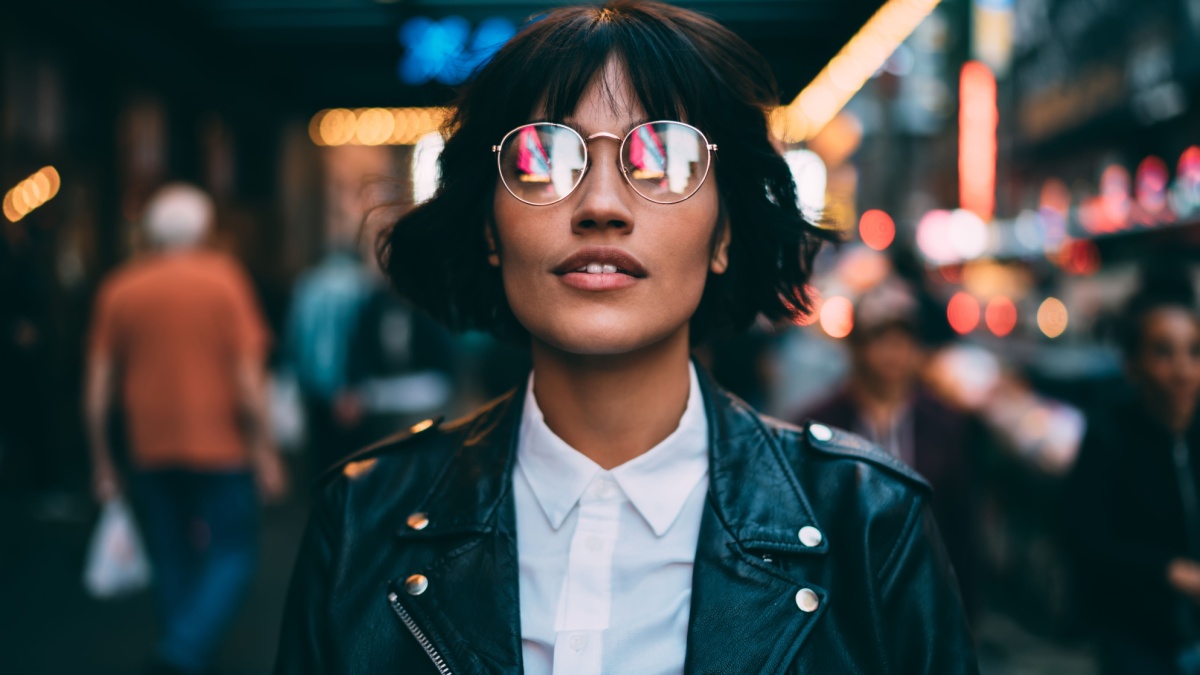Trendy dressed tourist in stylish eyeglasses with neon reflection looking up during evening sightseeing around metropolitan downtown, fashionable woman in spectacles hanging out in night city.