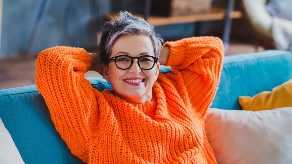 Photo of adorable satisfied aged person sit cozy couch beaming smile arms behind head pastime recreation house inside.