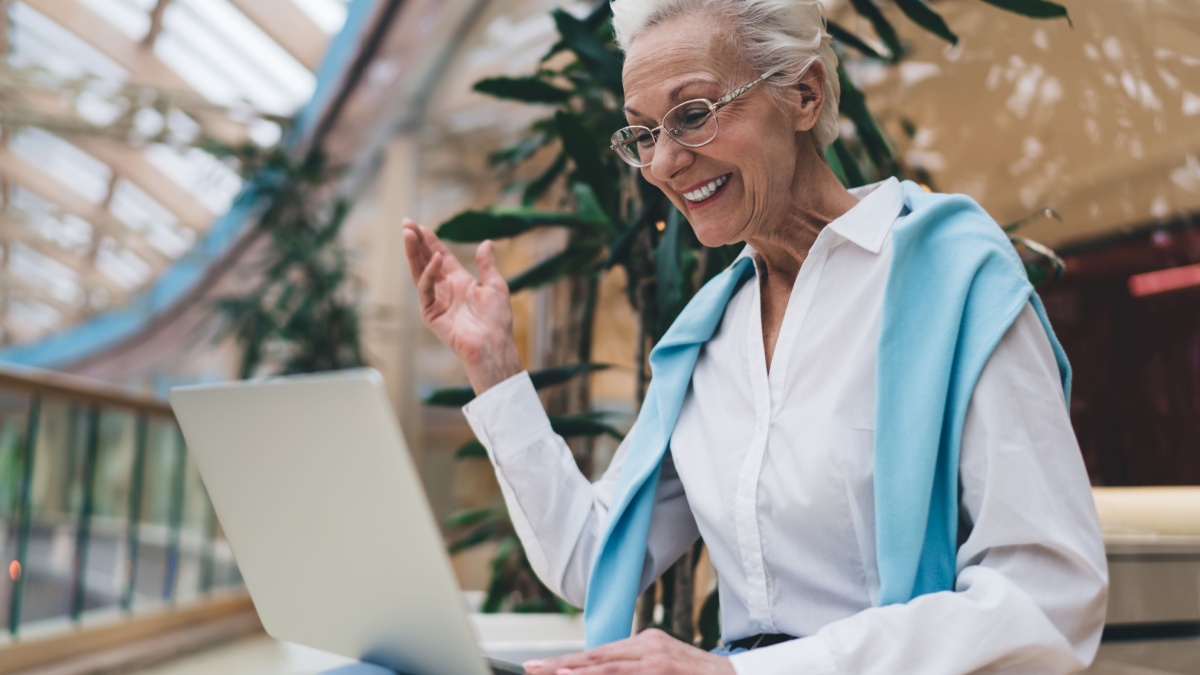 Elderly Caucasian woman in her 70s, smiling joyfully using laptop. She wear glasses, white shirt, , gesture with one hand, during video call.