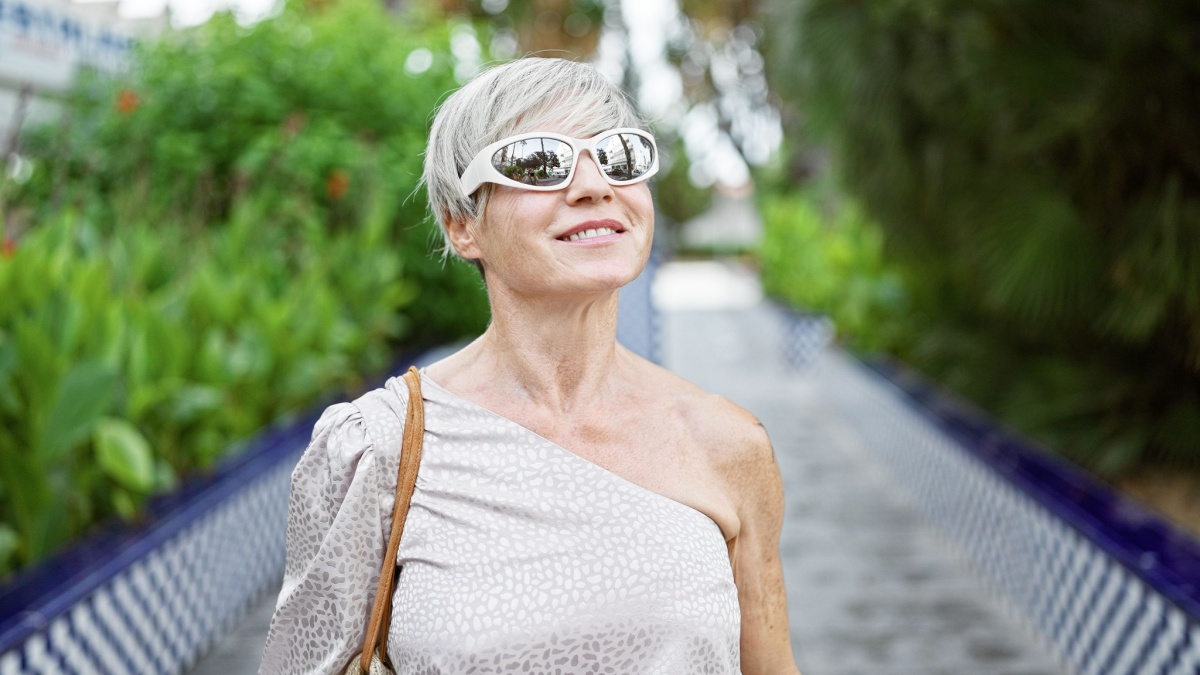 A confident mature woman enjoys a sunny day outdoors, surrounded by lush greenery in a serene park.
