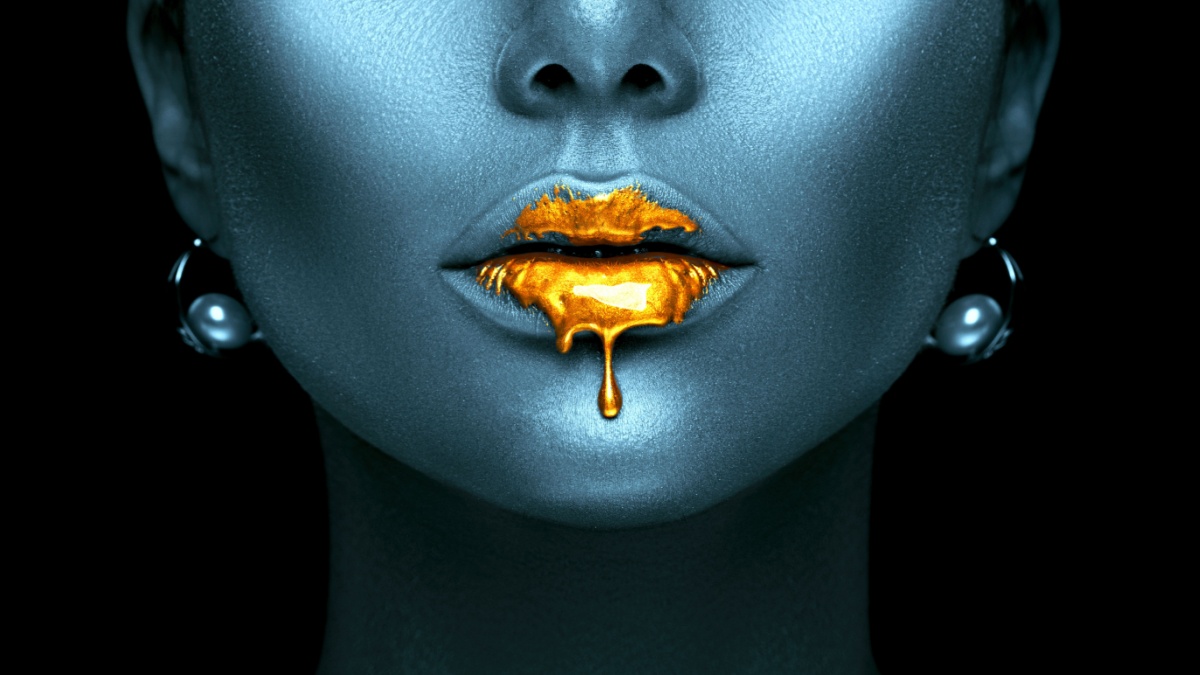 Gold Paint drips from the lips, lipgloss dripping from sexy lips, golden liquid drops on beautiful model girl's mouth, creative abstract dark blue skin make-up.